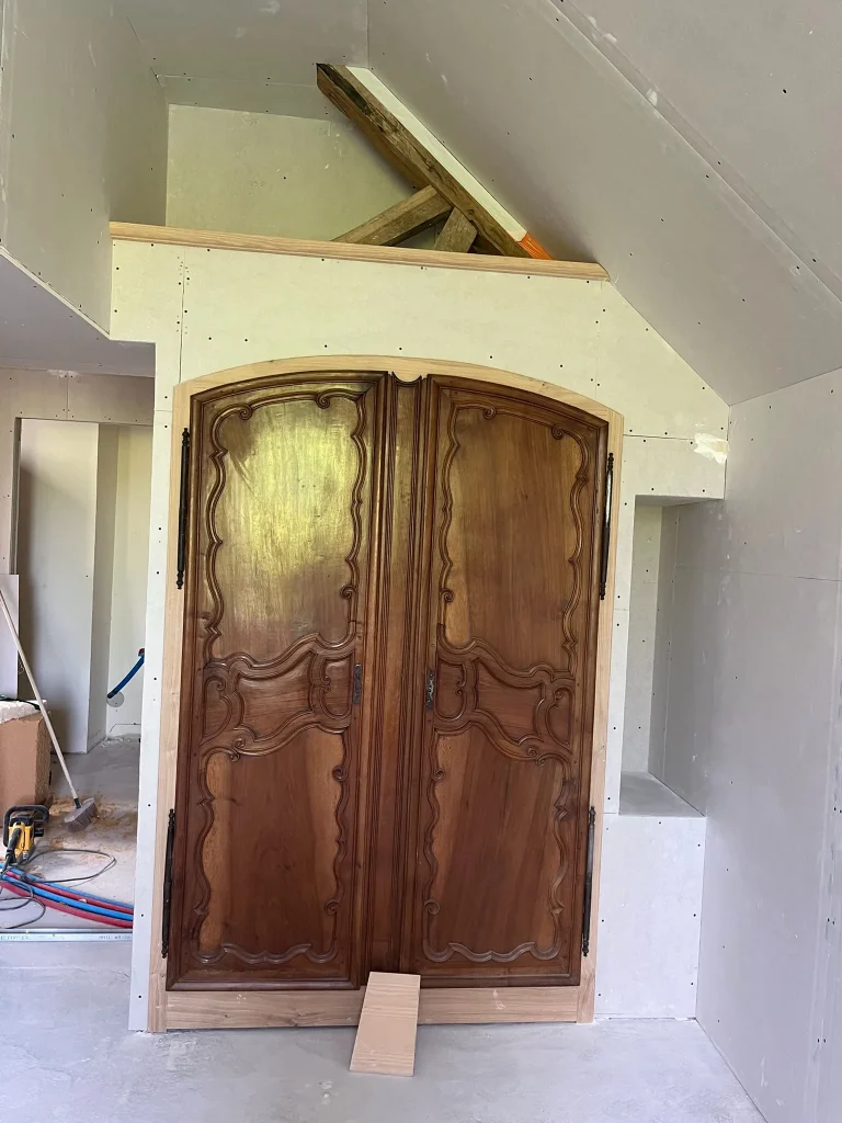 Plaques de plâtre ajustées autour d'un placard à portes en bois, sous un plafond en pente avec poutre apparente