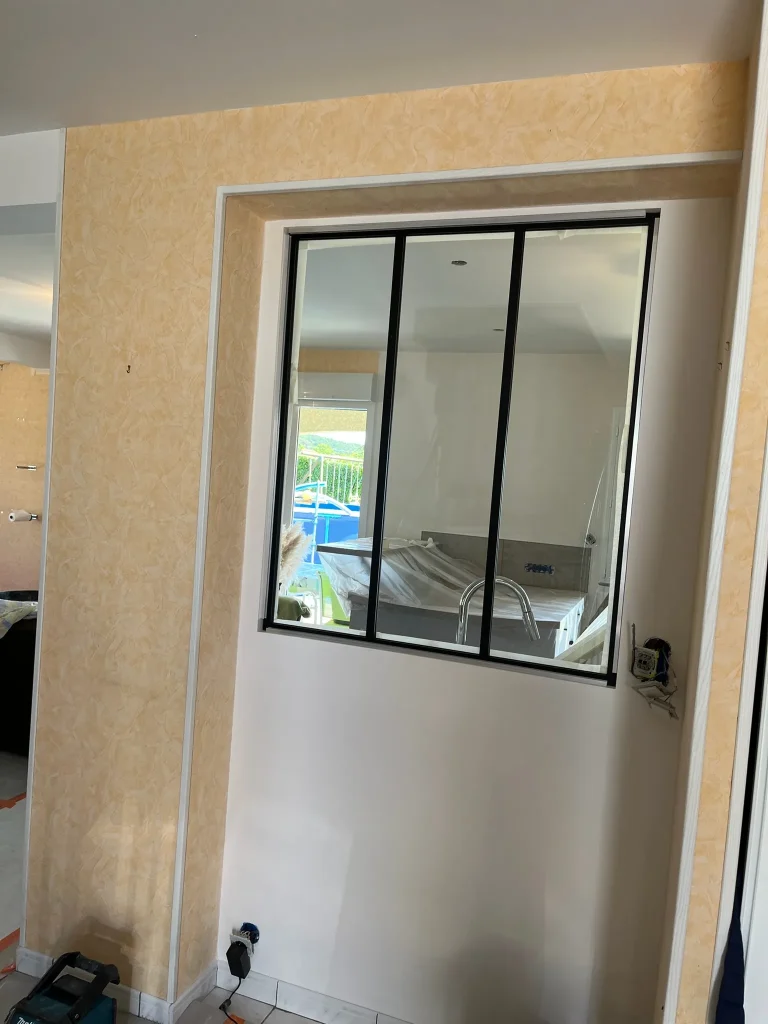 Mur habillé d'un revêtement beige texturé autour d'une verrière à cadre noir