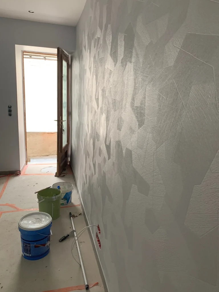 Mur habillé de papier peint gris à motifs géométriques, avec seau et rouleau au sol