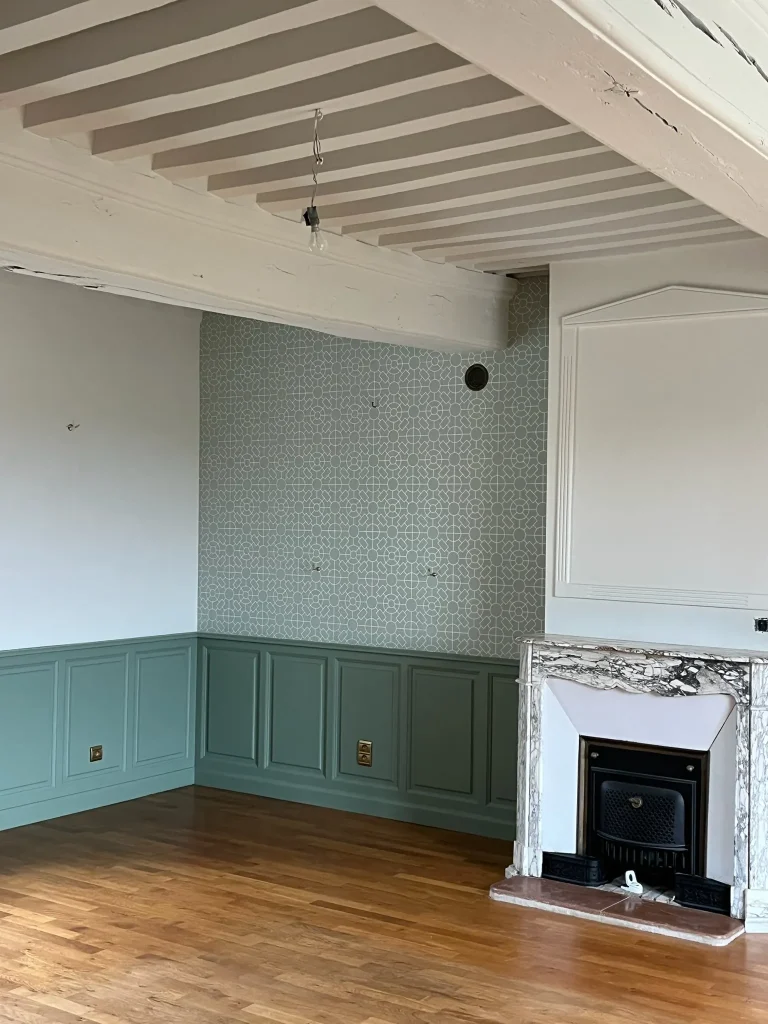 Pièce avec papier peint à motifs, soubassements verts, poutres peintes et cheminée