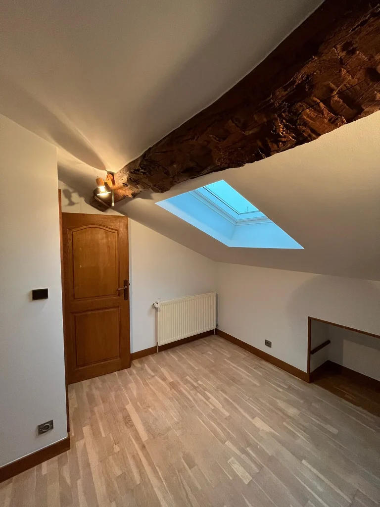 Pièce sous combles avec plafond blanc en pente, poutre apparente et fenêtre de toit