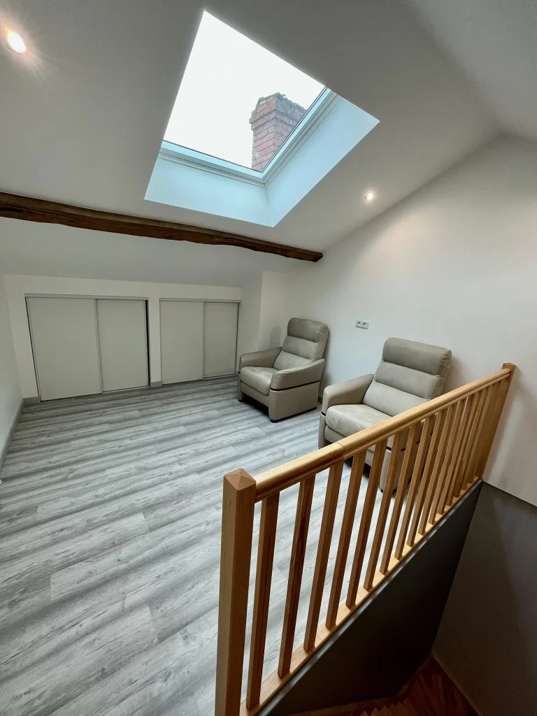 Pièce sous combles avec sol effet bois gris, deux fauteuils, fenêtre de toit et garde-corps en bois