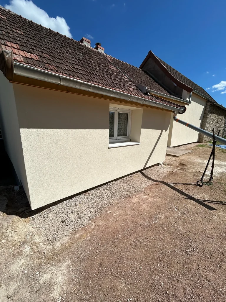 Façade claire enduite avec fenêtre sous une toiture en tuiles et sol protégé